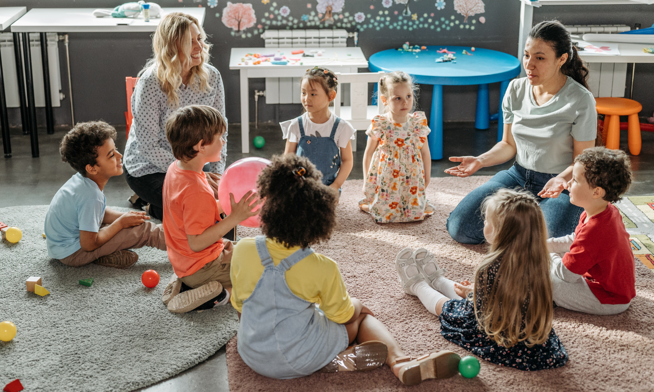 Dos maestras de Educación infantil sentadas en el suelo con varios niños y niñas